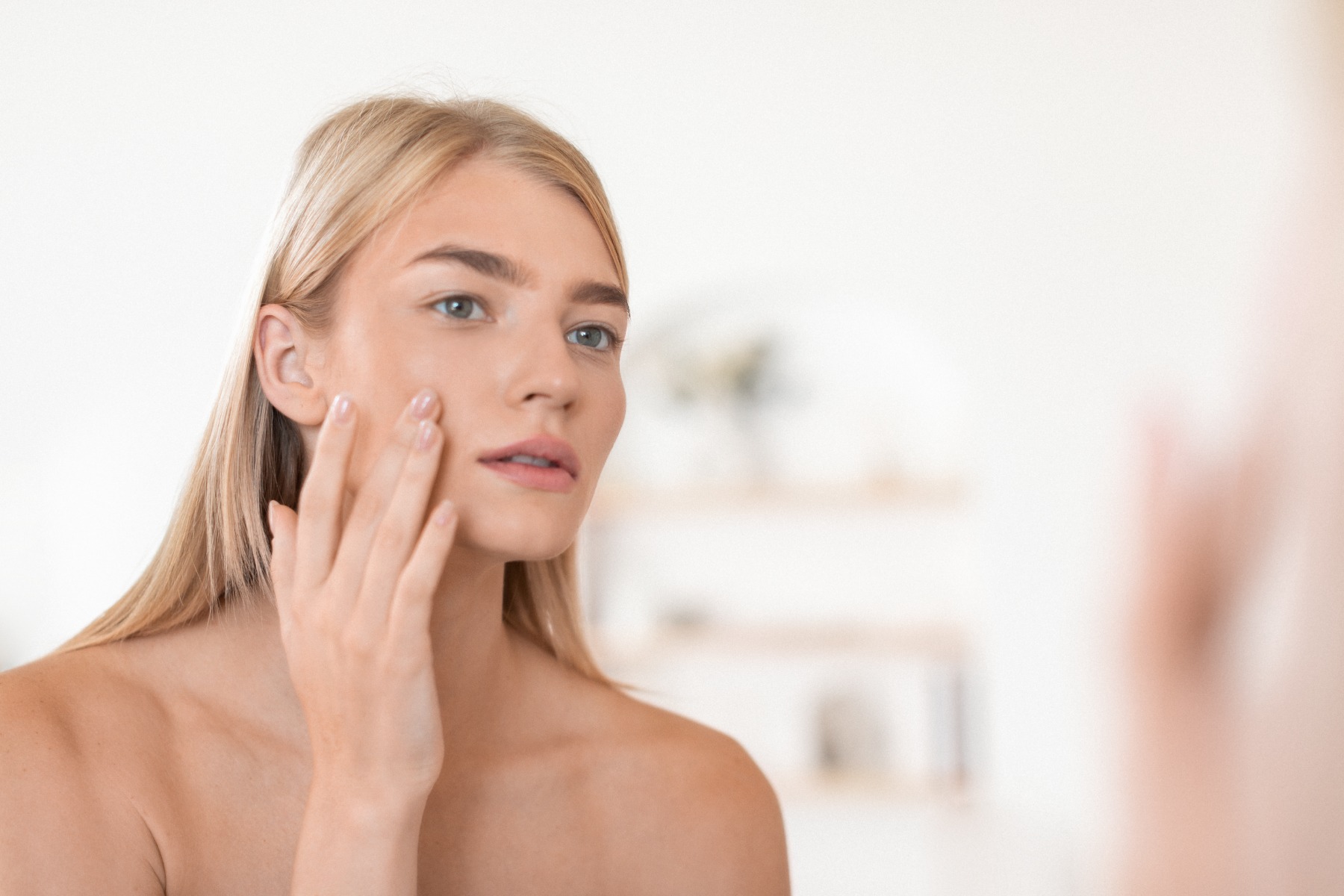 Frau mit langen, blonden Haaren betrachtet ihr Spiegelbild und berührt mit einer Hand sanft ihr Gesicht.